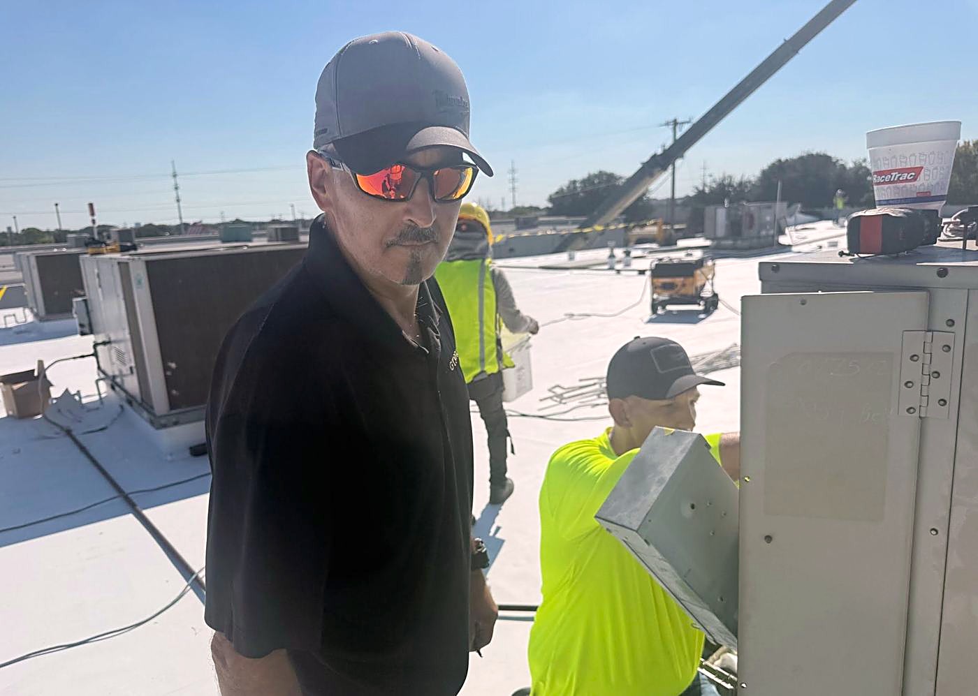 A man in a cap and sunglasses in the foreground on a white flat roof, with two workers and rooftop units behind him
