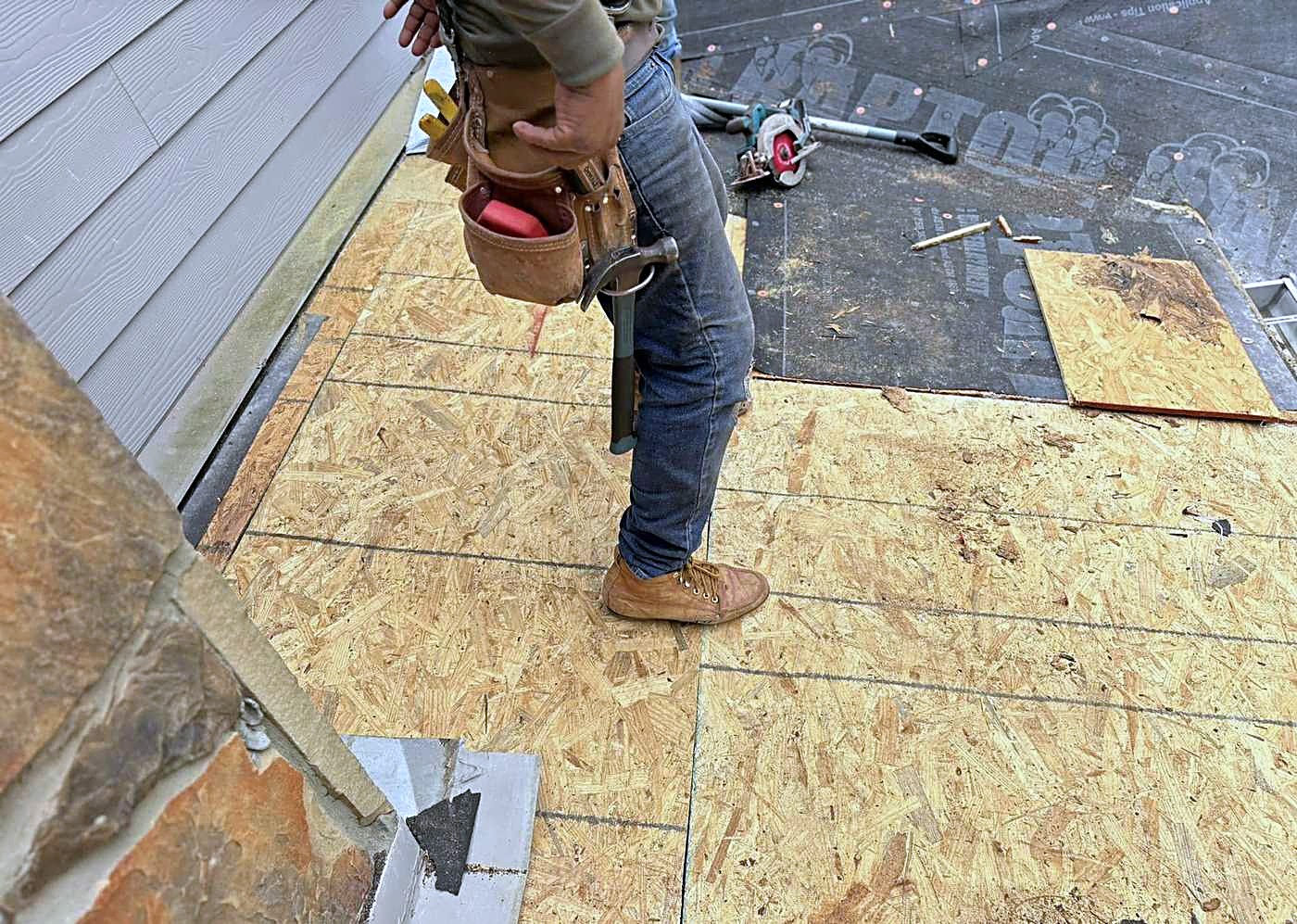 A roofer standing on fresh plywood decking beside a stone chimney