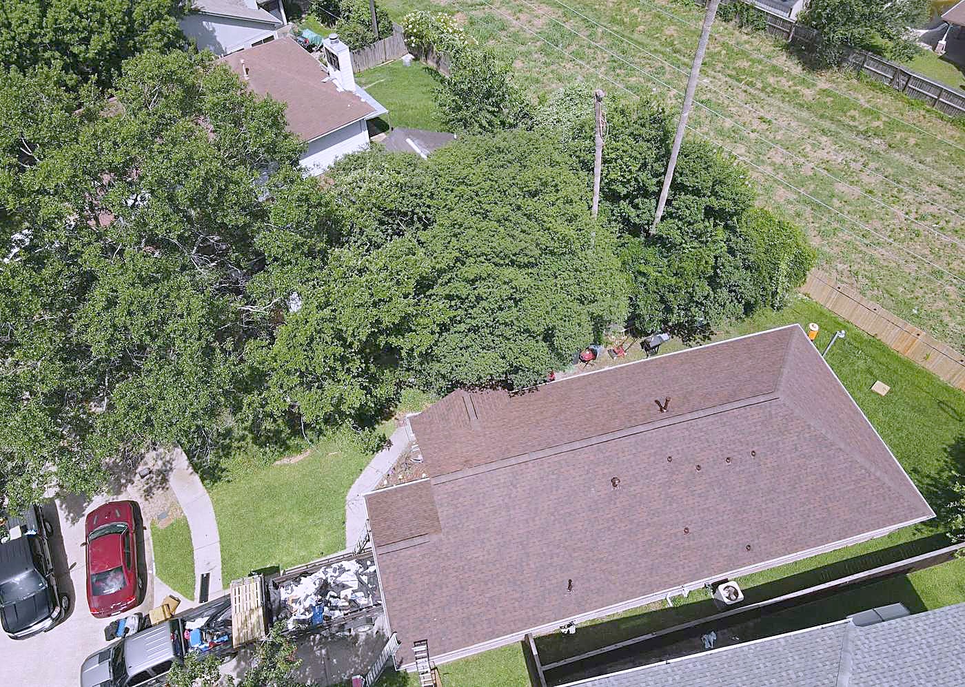 Aerial view of a finished brown roof beside a large tree canopy, with a tear-off debris pile on the driveway